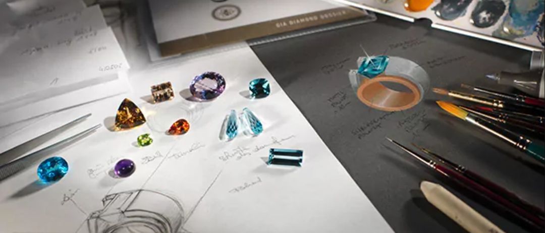 Eine Sammlung verschiedener Edelsteine und Schmuckdesign-Skizzen auf einem Schreibtisch, mit einem Buch mit dem Titel 'GIA Diamond Design' im Hintergrund.
