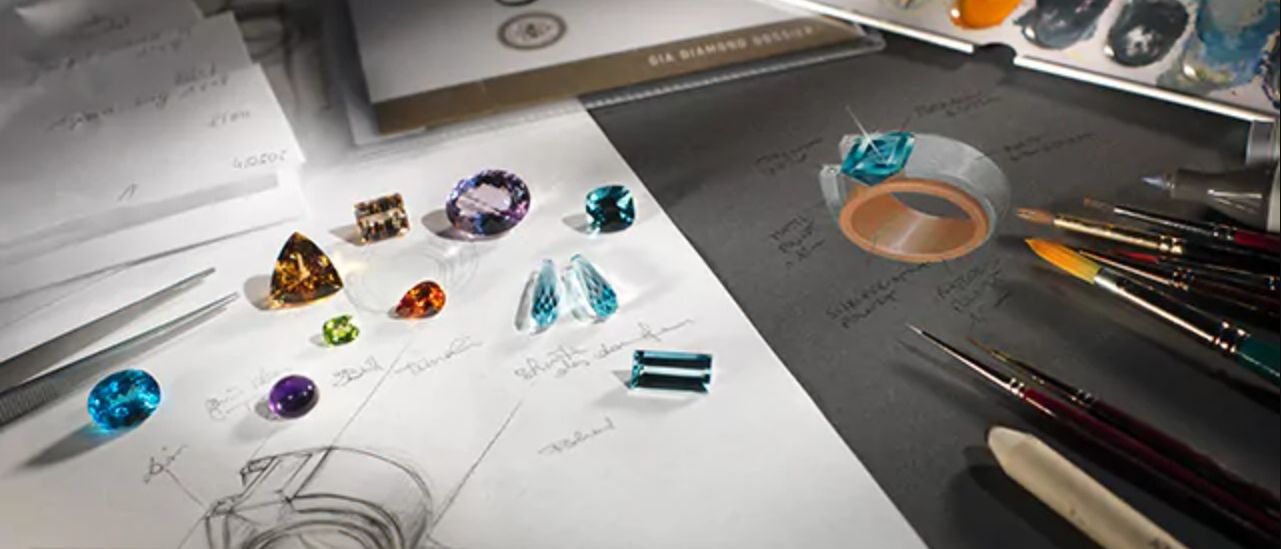 Eine Sammlung verschiedener Edelsteine und Schmuckdesign-Skizzen auf einem Schreibtisch, mit einem Buch mit dem Titel 'GIA Diamond Design' im Hintergrund.