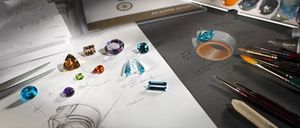 Eine Sammlung verschiedener Edelsteine und Schmuckdesign-Skizzen auf einem Schreibtisch, mit einem Buch mit dem Titel 'GIA Diamond Design' im Hintergrund.