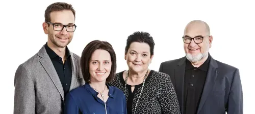 Vier Personen, zwei Männer und zwei Frauen, lächeln und posieren für ein Foto. Der Mann links trägt eine Brille und einen grauen Anzug, während die Frau links ein blaues Hemd trägt. Die Frau in der Mitte hat kurze Haare und eine Halskette, und der Mann rechts hat einen Bart und eine Brille.
