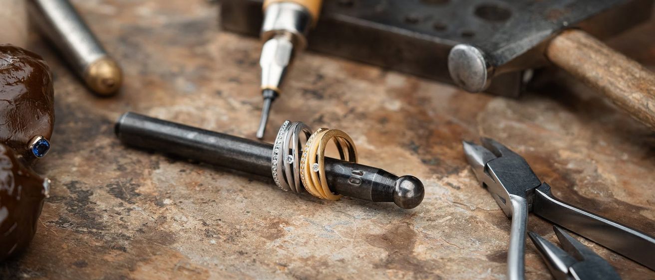 Ein Metallstab mit Ringen wird von einer Zange auf einer strukturierten Oberfläche gehalten. Eine Bohrmaschine und andere Werkzeuge sind in der Nähe.