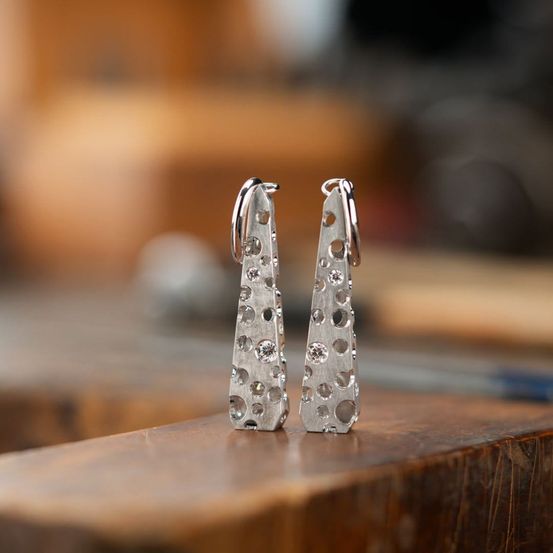 Zwei silberne Ohrringe mit geometrischem Muster und kleinen Diamanten liegen auf einer Holzoberfläche. Der Hintergrund ist unscharf.