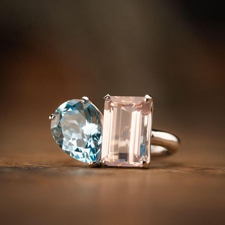Zwei Edelsteine sind auf einer hölzernen Oberfläche zu sehen. Der linke ist ein blauer Topas in Herzform, und der rechte ist ein rosa Turmalin. Sie sind beide an einem goldenen Ring befestigt.