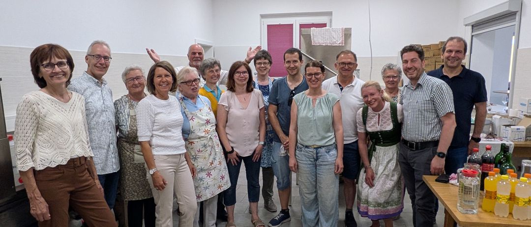 Eine Gruppe von Menschen posiert für ein Foto in einem Raum mit weißen Wänden und gefliesten Böden. Einige tragen Brillen, Uhren und Schuhe. Eine Frau in einem Kleid hat einen Gürtel. Ein Holztisch und eine blaue Kiste befinden sich rechts.
