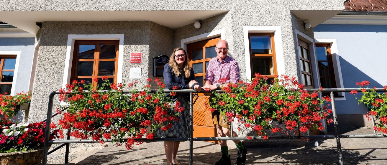 Zwei Personen stehen auf dem Balkon des Gasthofs Kraxner, lächeln und halten Händchen. Das Gebäude hat ein Schild mit der Aufschrift Gasthof Kraxner, mit Blumen und einer Tür.