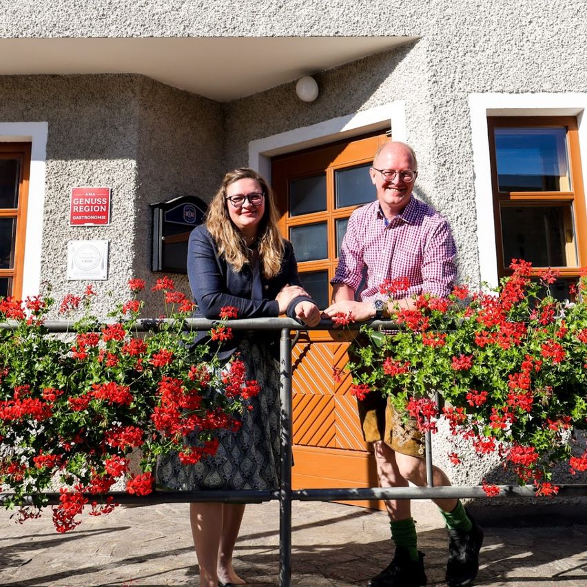 Two people stand on the balcony of Gasthof Kraxner, smiling and holding hands. The building has a sign reading Gasthof Kraxner, with flowers and a door.