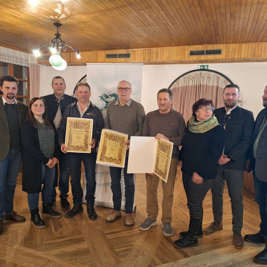 Eine Gruppe von Menschen in formeller Kleidung posiert mit Zertifikaten und Auszeichnungen in einem Raum mit Holzboden und Holzdecke.