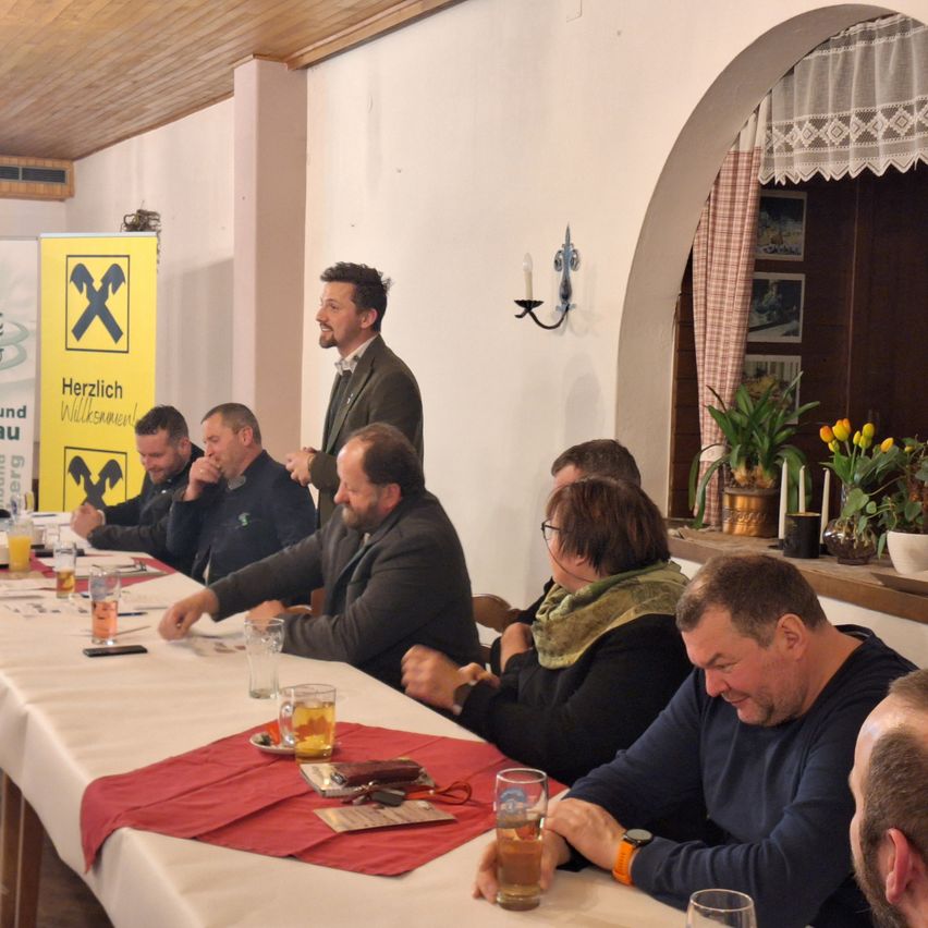 Mehrere Personen sitzen an einem langen Tisch, mit einem Mann, der vor ihnen steht. Der Tisch ist mit Gläsern, Tellern und verschiedenen Gegenständen gedeckt. Hinter ihnen befindet sich ein Fenster mit Vorhängen und Pflanzen.