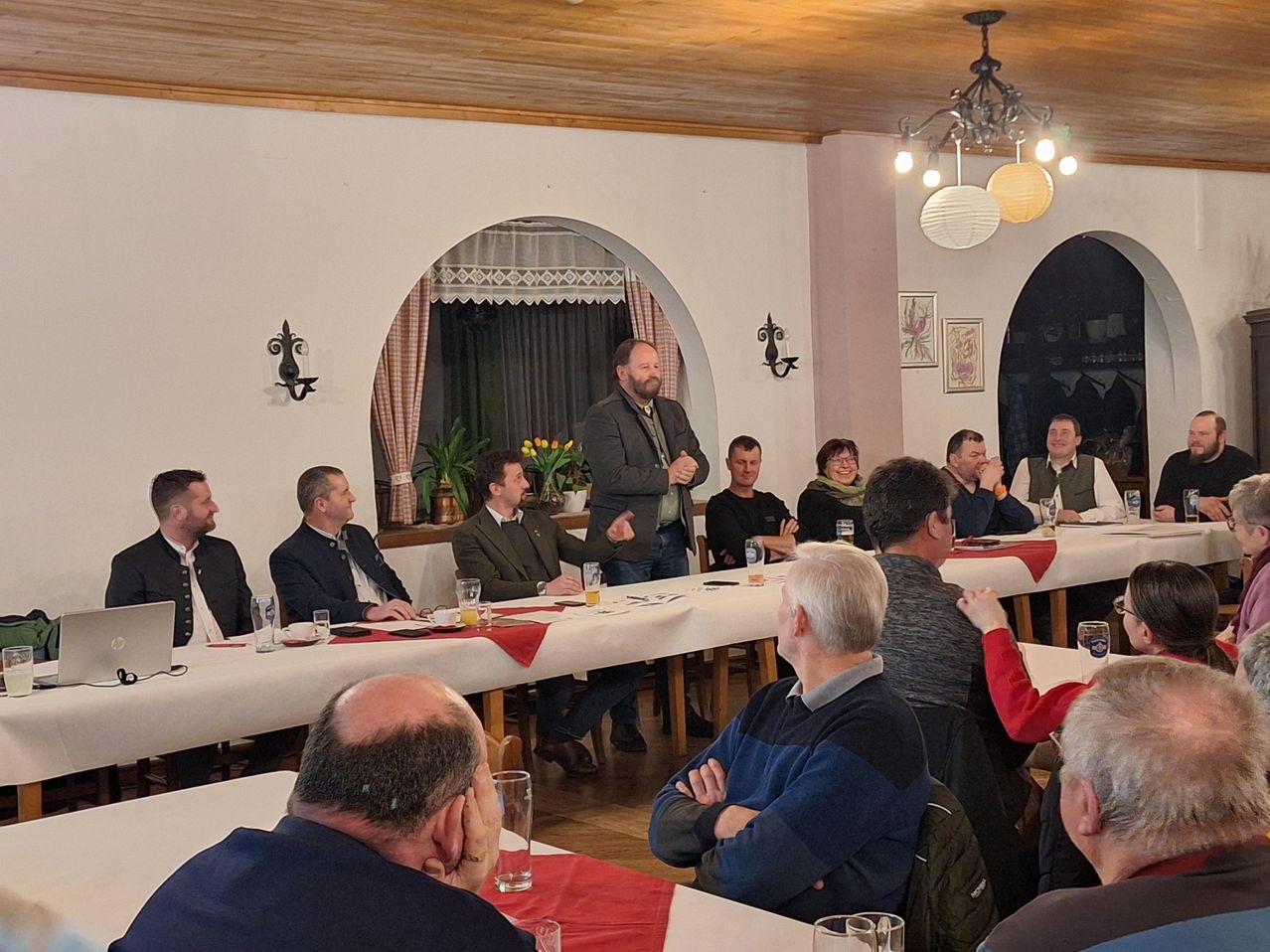 Eine Gruppe von Männern sitzt an einem Tisch in einem Raum, möglicherweise für eine Besprechung. Sie tragen formelle Kleidung, und einige von ihnen halten Gläser. Der Mann, der vor ihnen steht, spricht wahrscheinlich. Der Raum hat eine Holzdecke und Wände mit einer hängenden Lampe. Es gibt auch Bilderrahmen an der Wand.