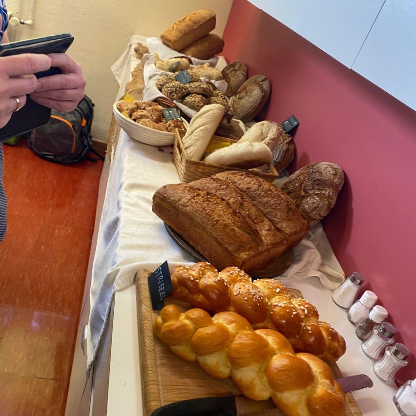 A table with a variety of breads including loaves, challah, and others on a white tablecloth. A person holds a smartphone.