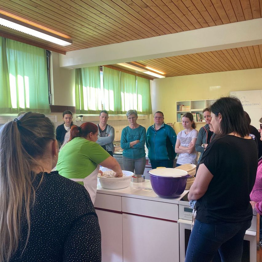 A woman is preparing food in a kitchen with several people watching. The kitchen has a counter, a sink, and a wooden ceiling. A whiteboard is mounted on the wall.
