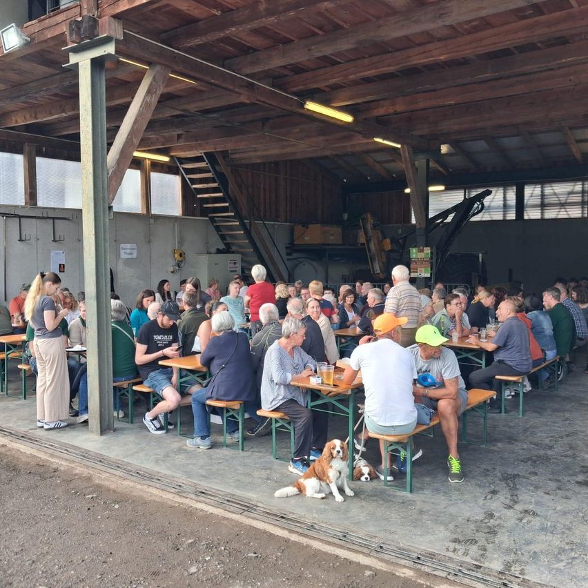 Eine große Gruppe von Menschen versammelt sich in einem überdachten Außenbereich. Holztische und -bänke sind in Reihen angeordnet. Das Dach wird von Holzbalken und Metallpfosten gestützt.