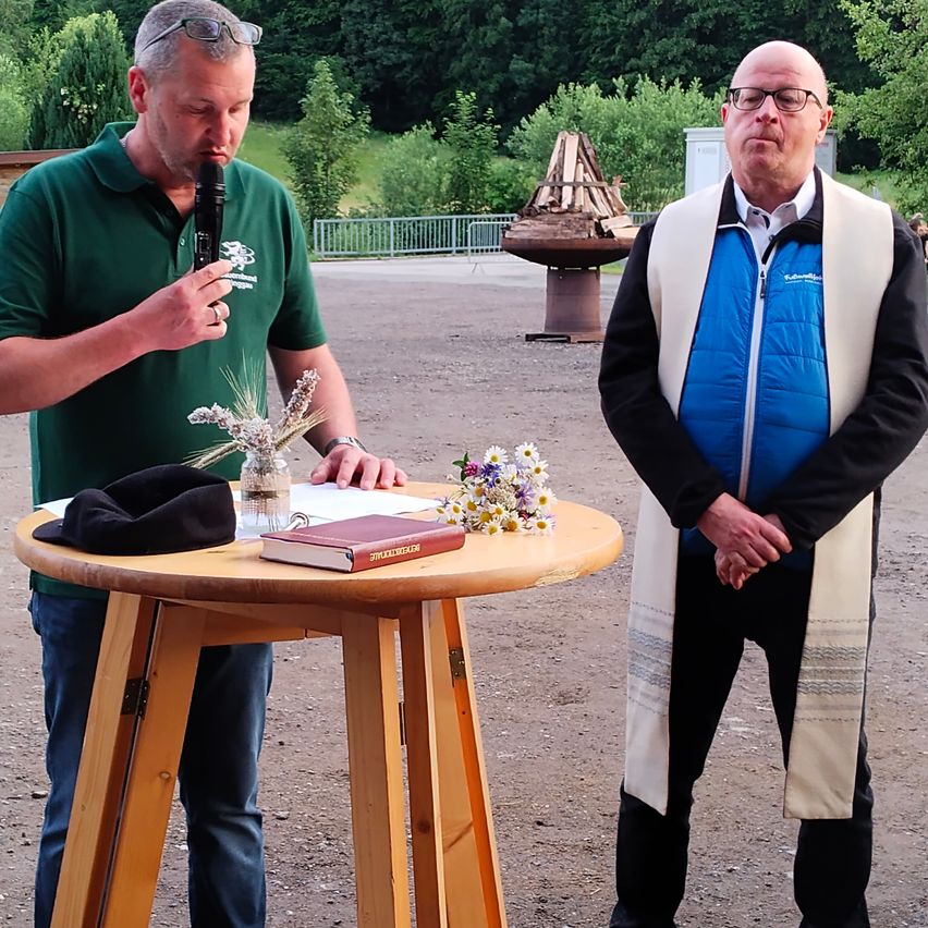 Zwei Männer stehen in einem Außenbereich. Einer hält ein Mikrofon und spricht, während der andere zuhört. Ein Holztisch mit einem Buch, Blumen und einem schwarzen Hut steht in der Nähe. Dahinter ist ein hölzerner Feuerkorb zu sehen.