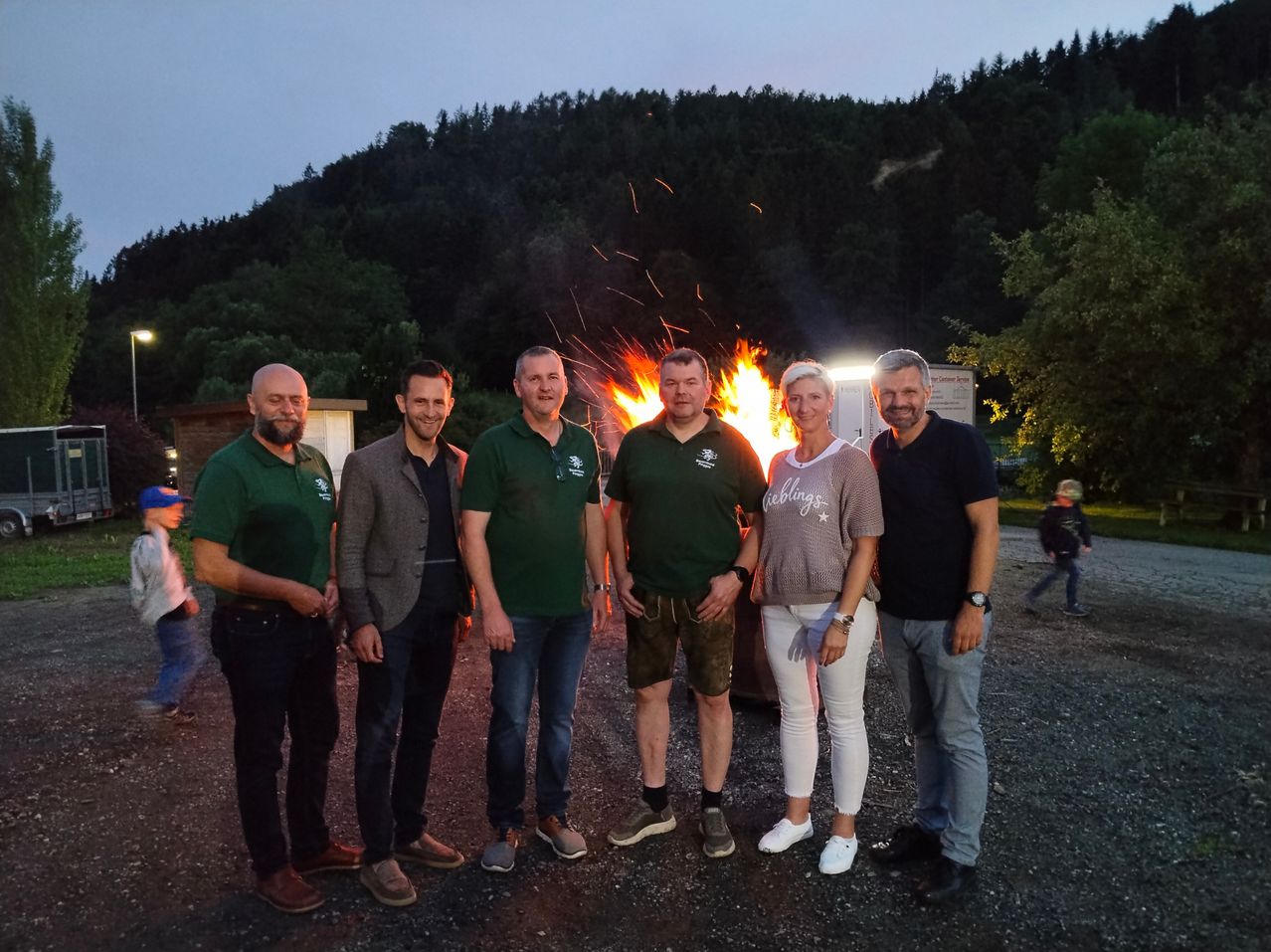 Fünf Erwachsene stehen vor einem Feuer in einer Outdoor-Umgebung. Dahinter befindet sich ein Wald.