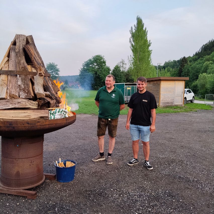 Zwei Männer stehen neben einem Feuerkorb mit brennendem Holz, umgeben von einer Kiesfläche. Im Hintergrund sind Bäume und ein kleines Gebäude zu sehen.