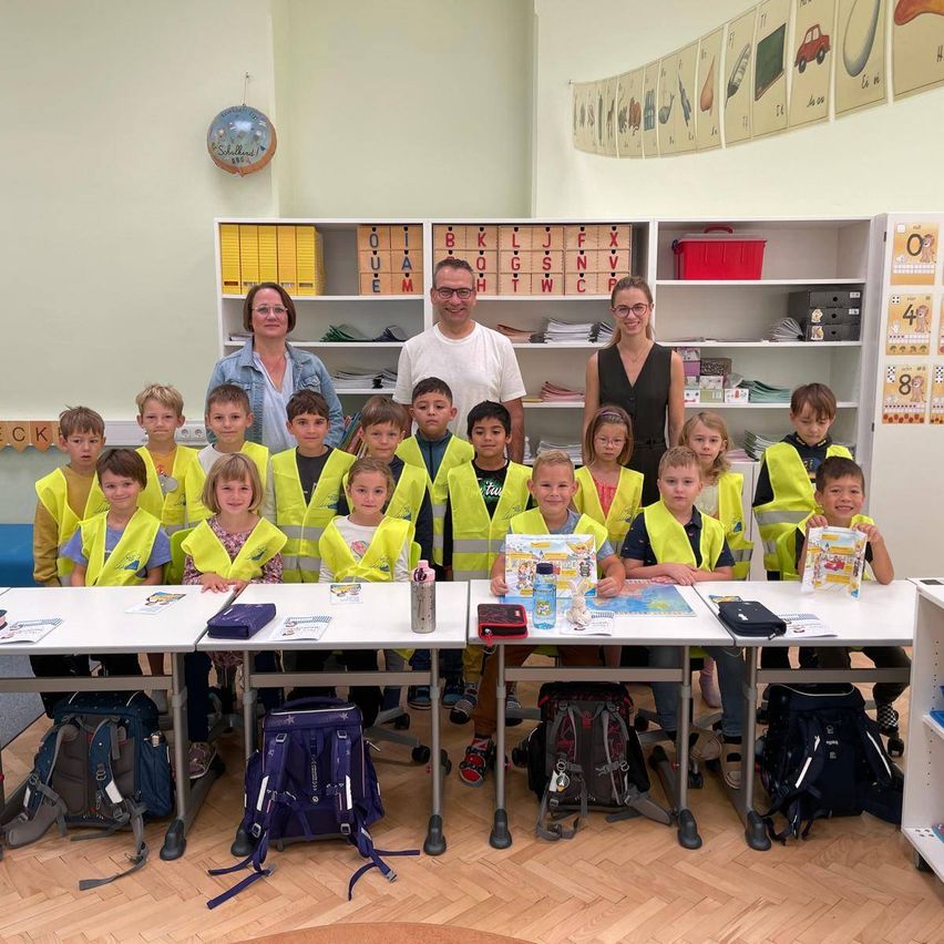 Eine Gruppe von Kindern in gelben Westen sitzt an Schreibtischen in einem Klassenzimmer und lächelt für ein Foto mit zwei Erwachsenen.