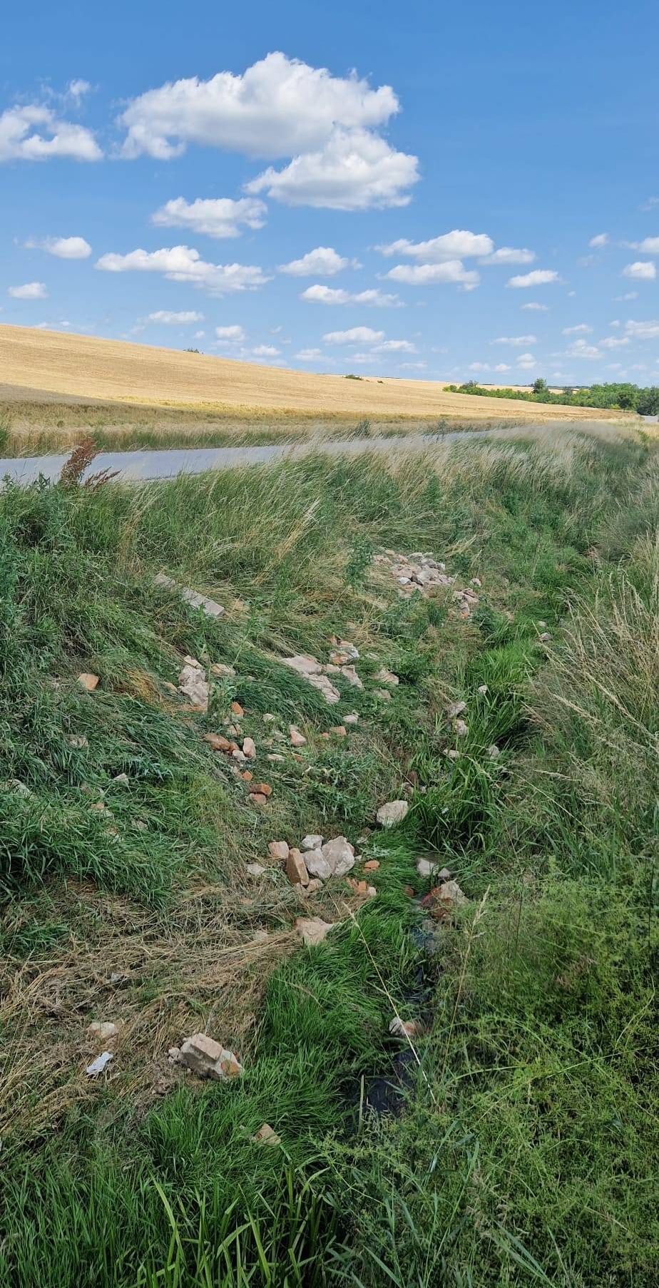Eine ländliche Landschaft mit einem schmalen Wasserlauf, der ein Grasfeld von einem goldenen Weizenfeld trennt, unter einem blauen Himmel mit verstreuten Wolken.