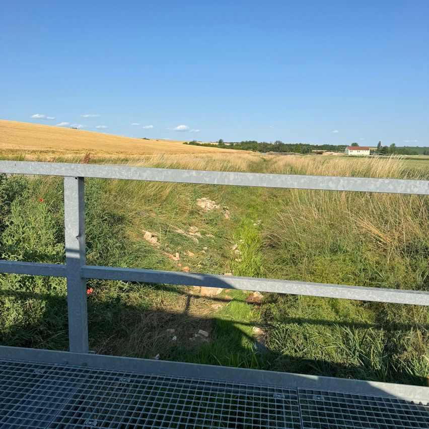 Eine erhöhte Ansicht eines Grasfeldes mit einem schmalen Pfad. Ein Metallgeländer ist vorhanden. In der Ferne befindet sich ein abgeerntetes Weizenfeld und ein Haus. Der Himmel ist klar mit einigen Wolken.