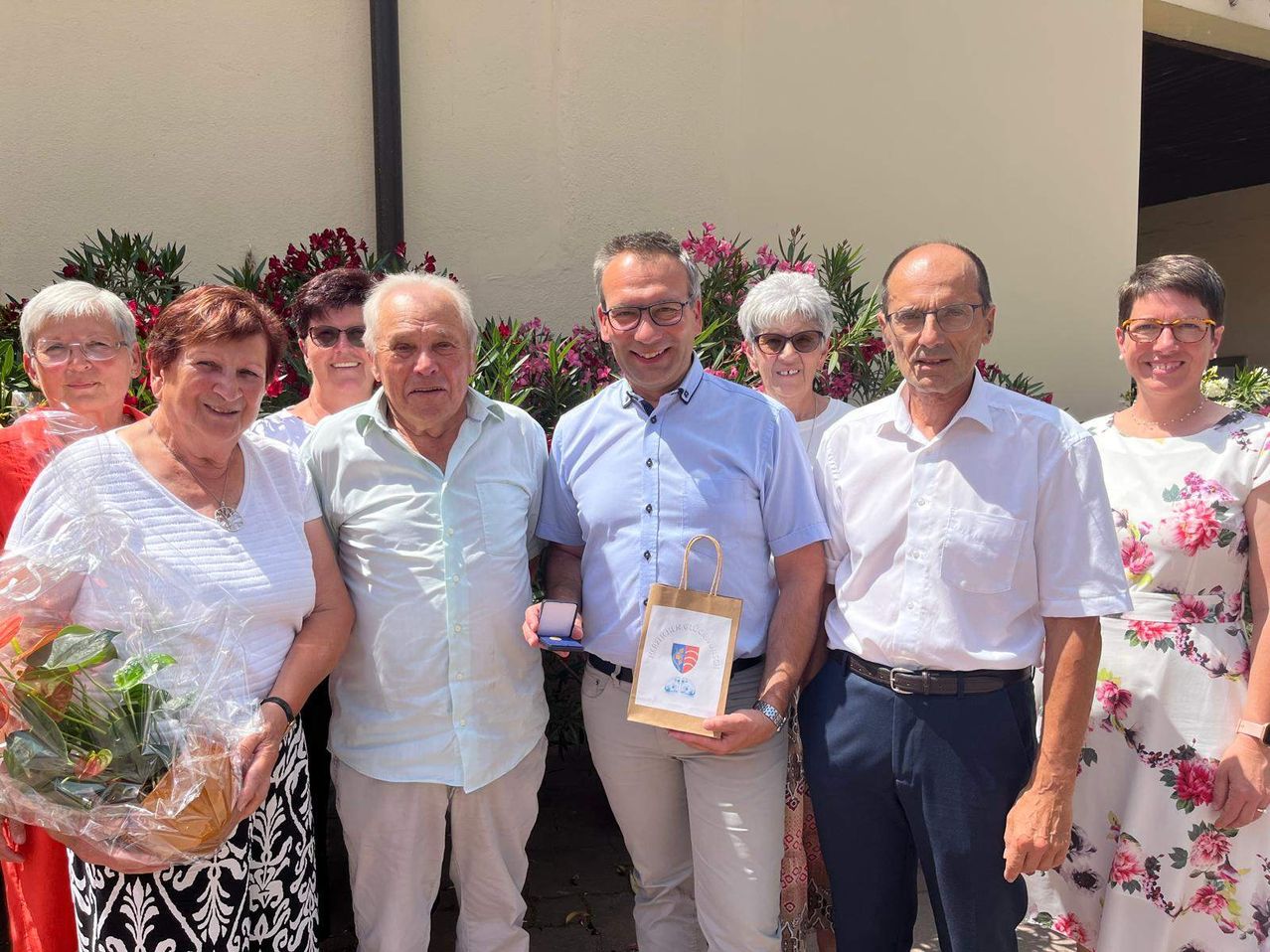 Sechs Menschen stehen vor einem Gebäude und lächeln für ein Foto. Der Mann in der Mitte hält einen Papierbeutel und eine Medaille. Die Frauen auf beiden Seiten halten Geschenke. Dahinter sind Blumen und eine Wand.