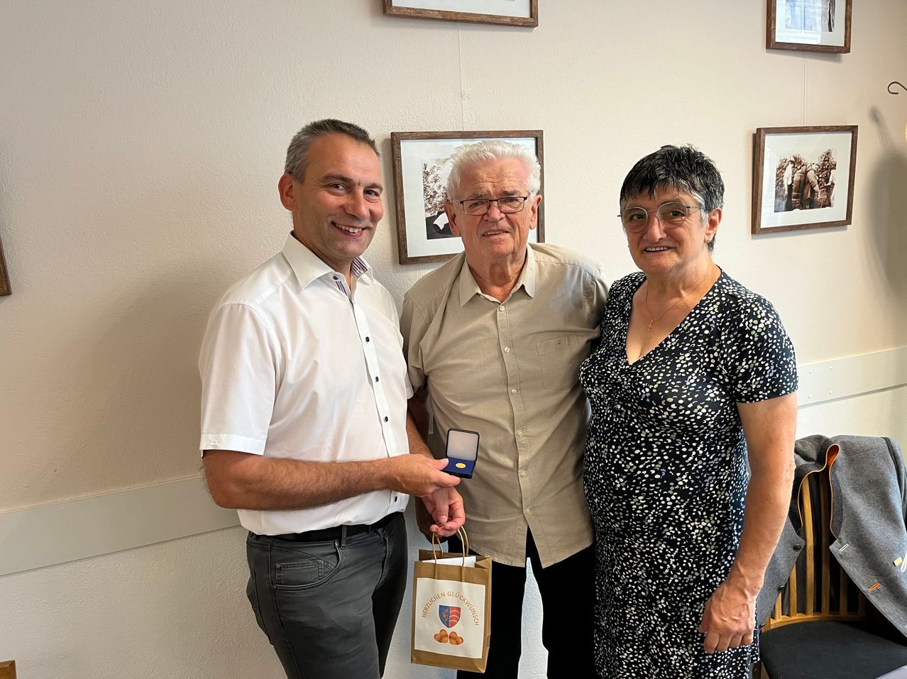 Drei Menschen stehen in einem Raum. Der Mann auf der linken Seite hält eine Geschenktüte. Der Mann in der Mitte hält eine Medaille. Die Frau auf der rechten Seite lächelt. Hinter ihnen hängen drei gerahmte Bilder an der Wand.
