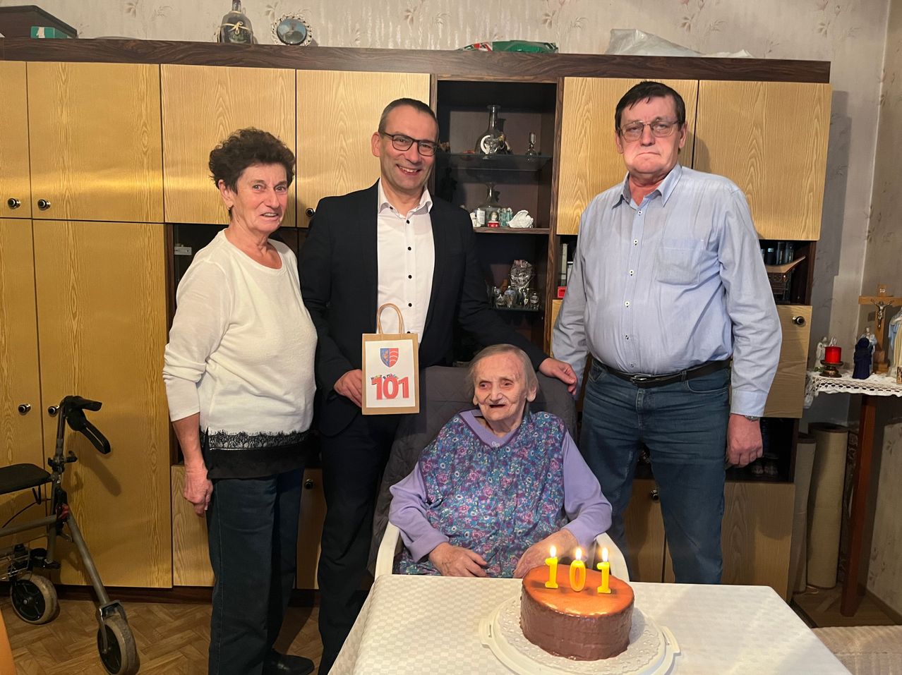 Vier Personen stehen um einen Tisch mit einem Geburtstagskuchen. Eine ältere Frau sitzt auf einem Stuhl und lächelt. Der Kuchen hat die Nummer 101 darauf. Dahinter steht ein Holzschrank mit verschiedenen Gegenständen.