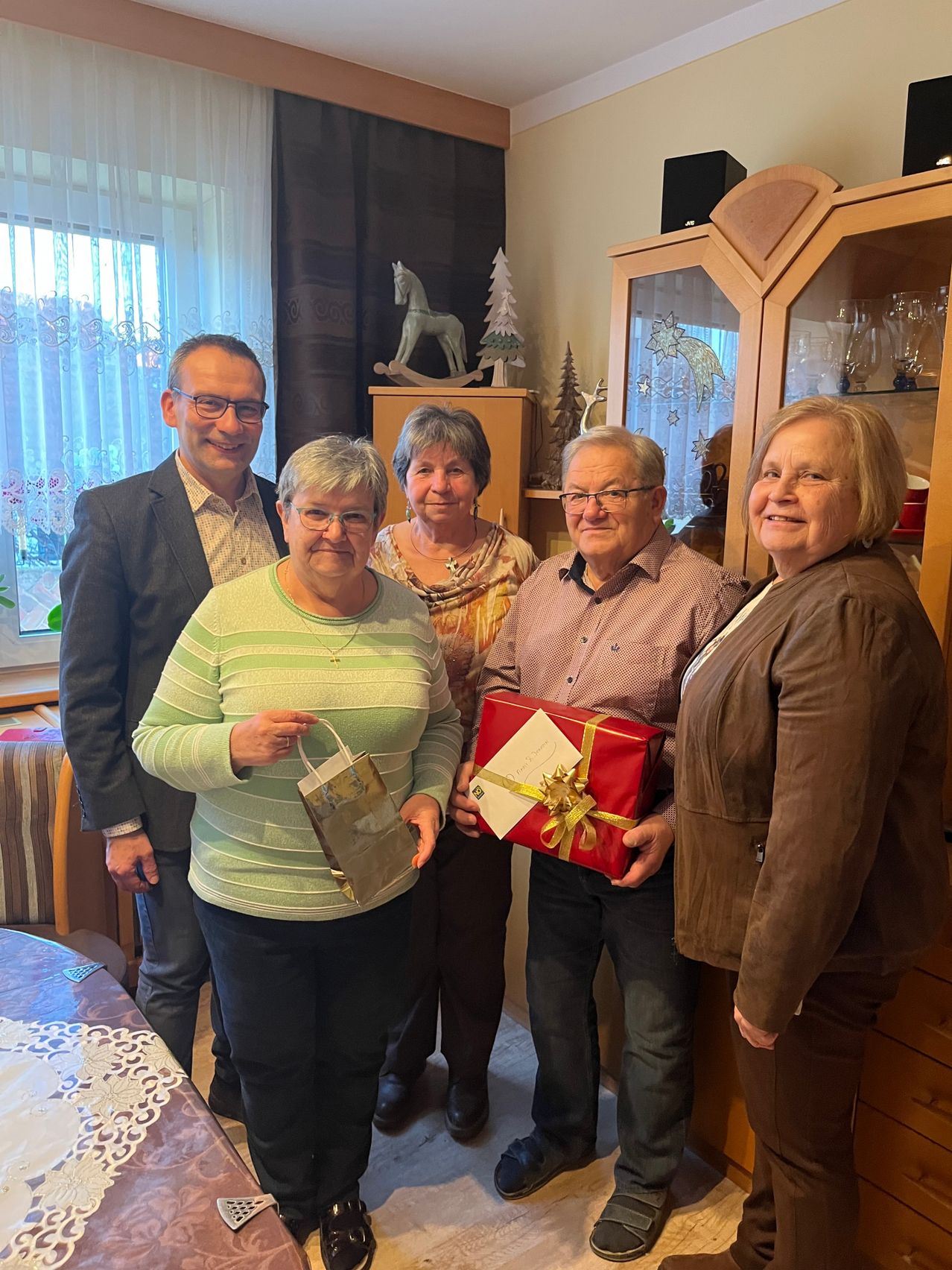 Fünf Menschen stehen in einem Raum mit festlicher Atmosphäre. Zwei Frauen und ein Mann halten Geschenktüten und ein eingewickeltes Geschenk. Die Frau auf der rechten Seite trägt eine braune Jacke. Hinter ihnen steht ein Schrank mit einem dekorativen Weihnachtsbaum und anderen Gegenständen.