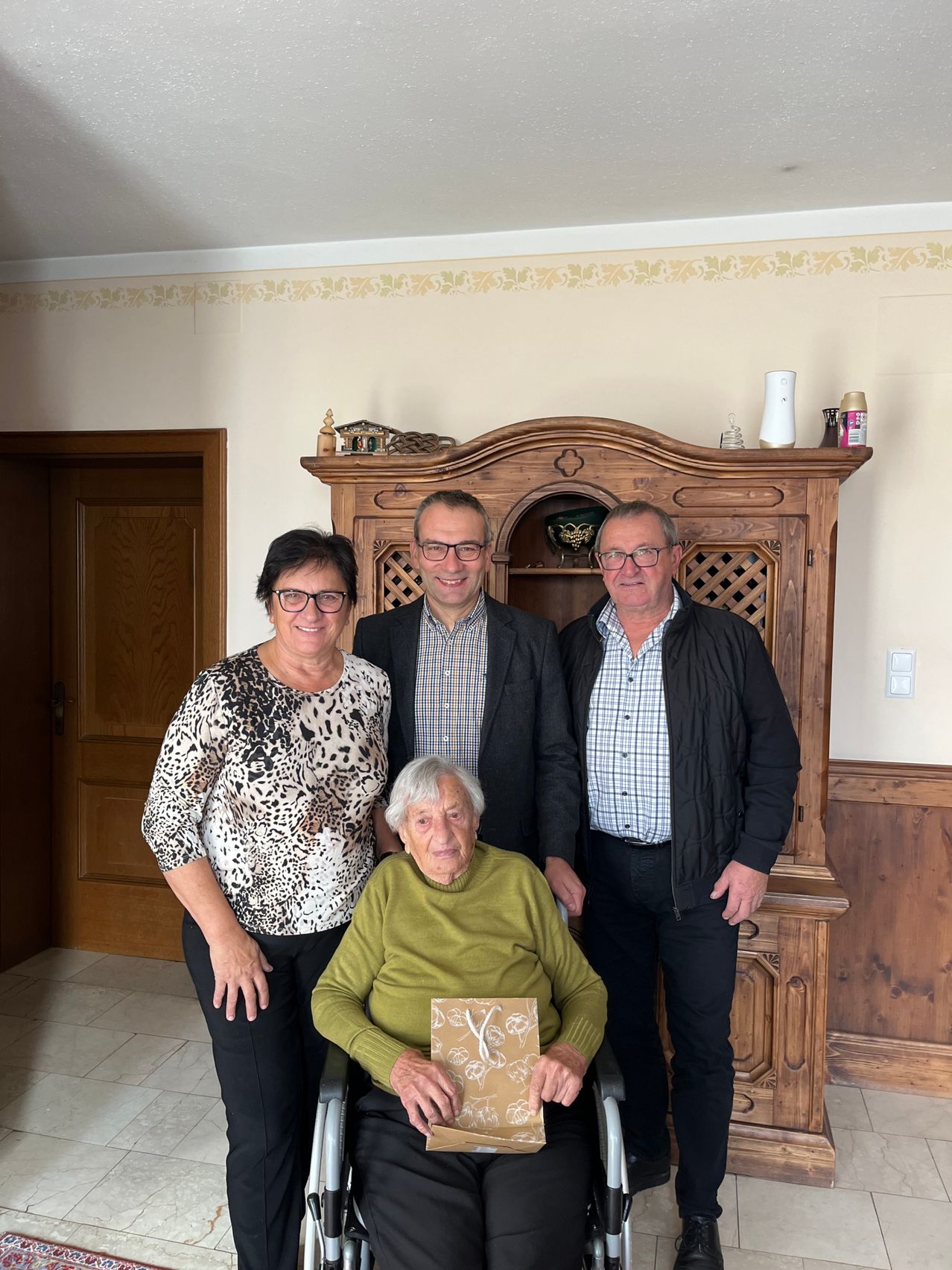 Vier Personen stehen in einem Wohnzimmer, wobei eine Frau im Rollstuhl ein eingewickeltes Geschenk hält. Dahinter steht ein Holzschrank mit verschiedenen Gegenständen darauf.