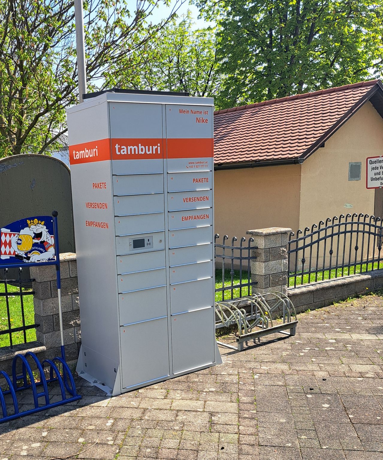 Ein großer grauer Postschließfach mit 'tamburi' darauf. Dahinter steht ein Gebäude mit einem braunen Dach. In der Nähe ist ein Fahrradständer.