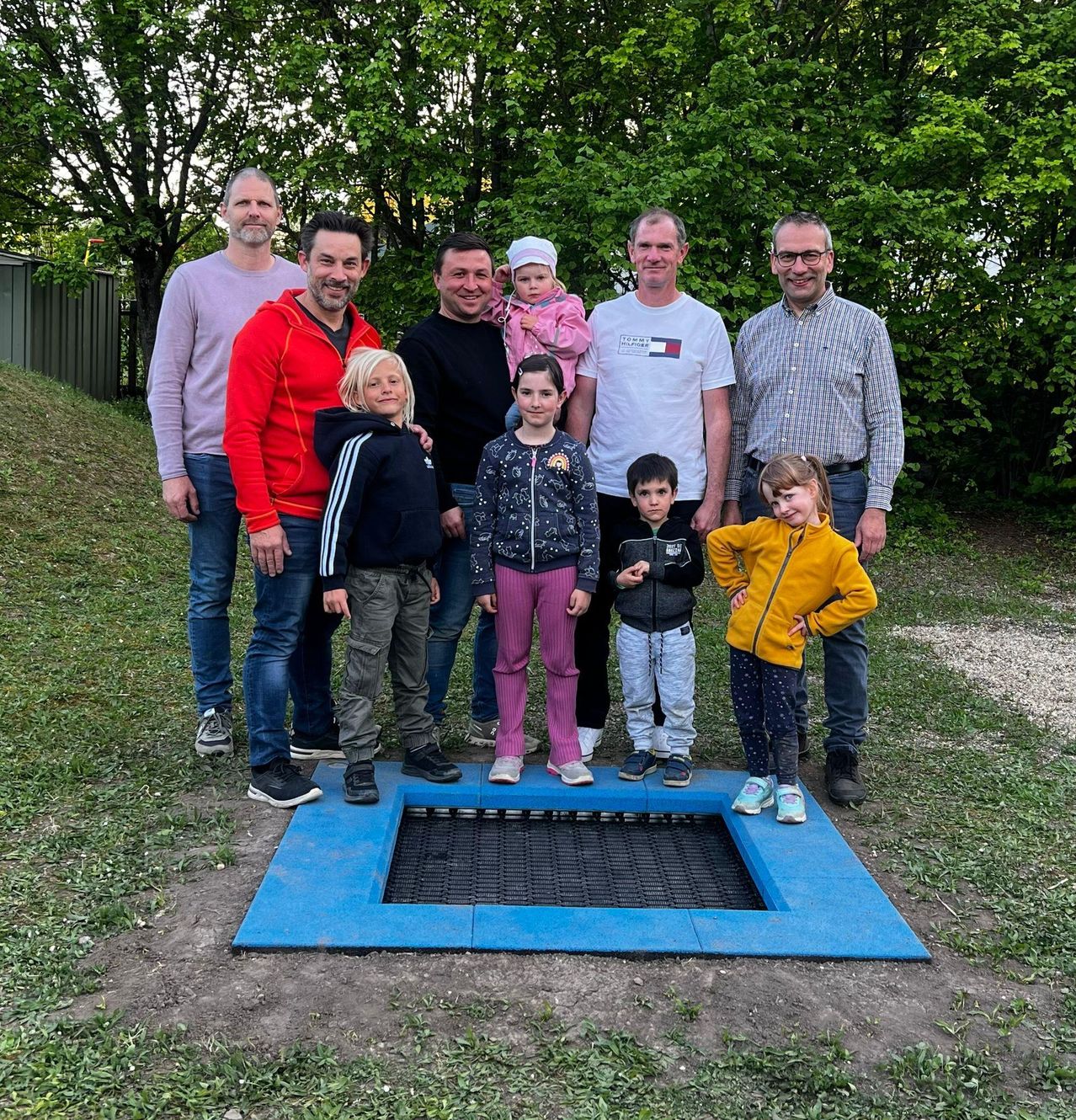 Eine Gruppe von Menschen, darunter Kinder, steht zusammen in einem Park in der Nähe einer blauen quadratischen Plattform.