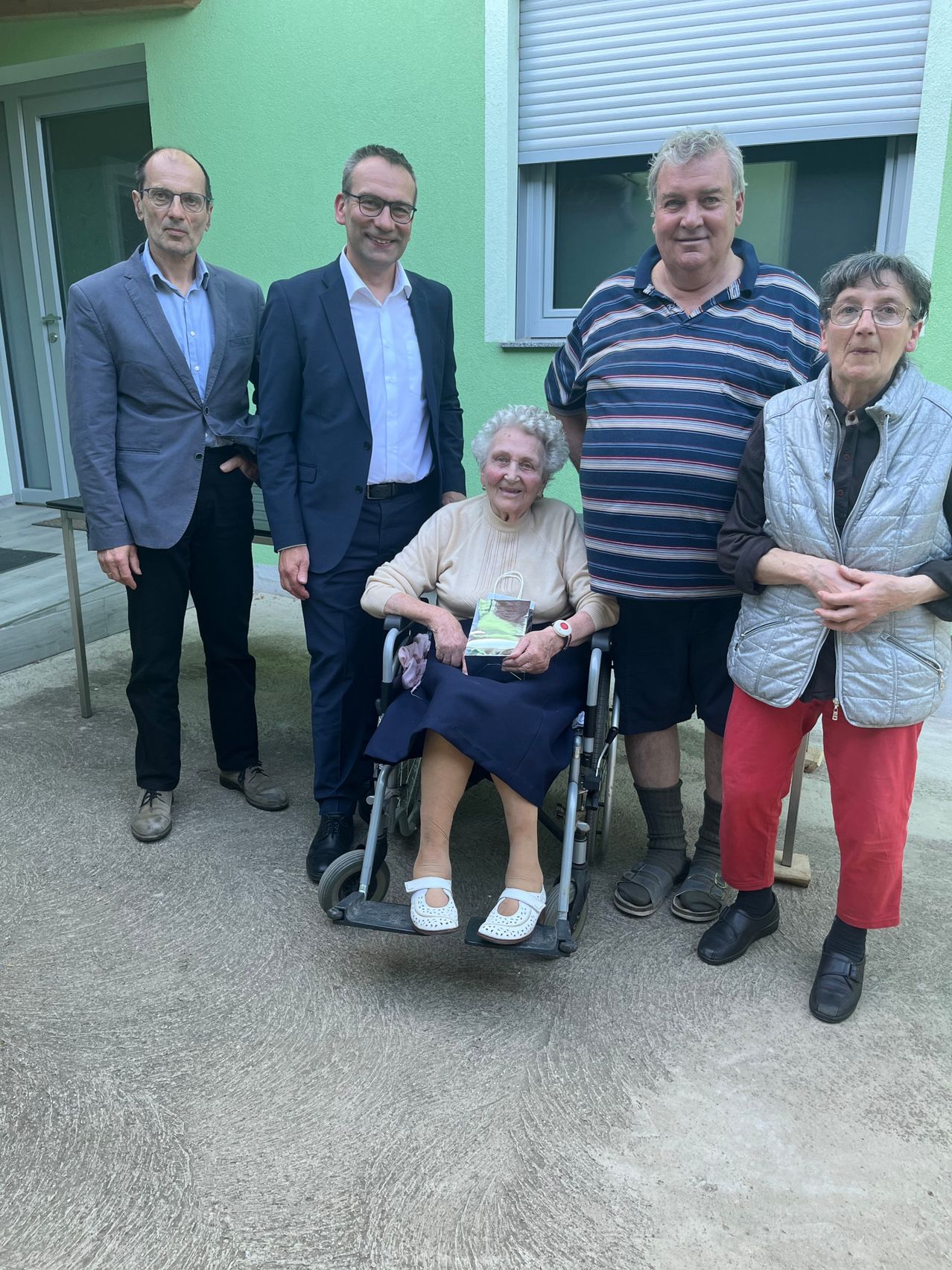 Five adults, three men and two women, are standing outside a house, smiling. The elderly woman in a wheelchair holds a book. The man in the middle wears a striped shirt.
