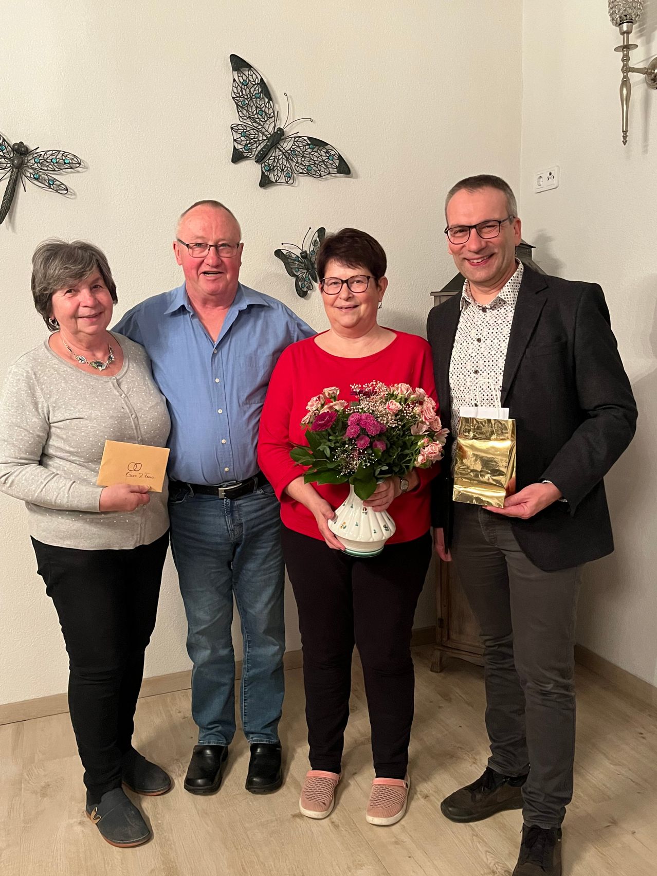 Vier Menschen stehen eng beieinander und lächeln. Eine Frau hält einen Blumenstrauß und eine andere Frau einen gelben Papierbeutel. Der Mann links trägt ein blaues Hemd und der Mann rechts einen schwarzen Anzug. Eine weiße Wand mit Schmetterlingsdekorationen steht hinter ihnen.