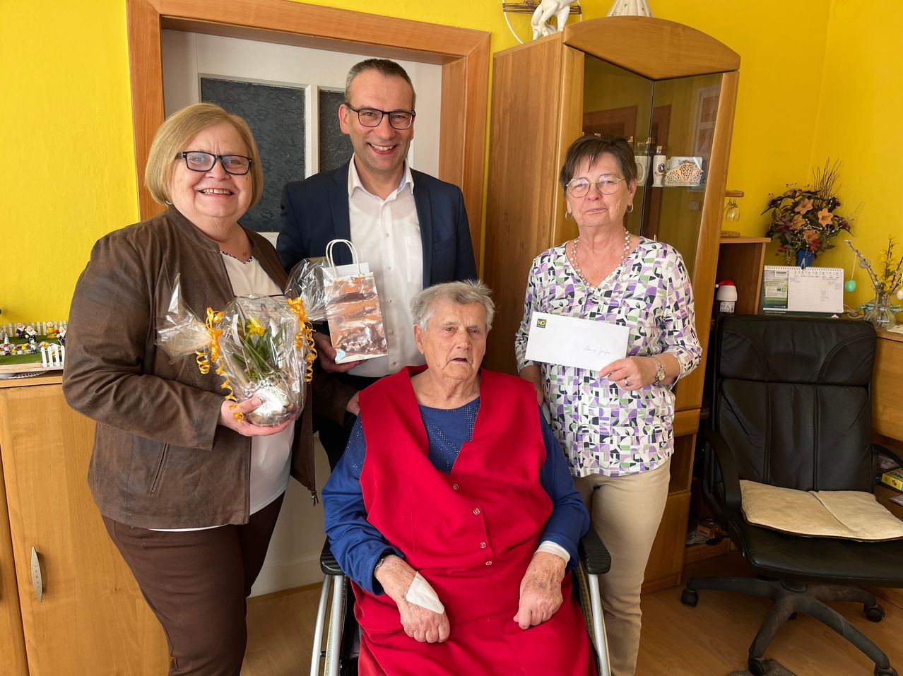Vier Personen stehen in einem Raum. Die Frau im Rollstuhl hält ein Papier und einen Blumenstrauß. Eine weitere Frau hält einen Blumenstrauß. Ein Mann hält einen Geschenkbeutel.