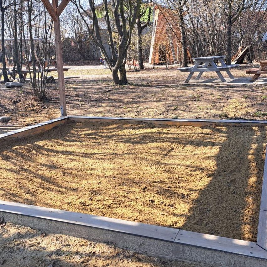 Ein Spielplatz im Freien mit einem Sandkasten, umgeben von Picknicktischen und Bäumen in einer Lichtung.