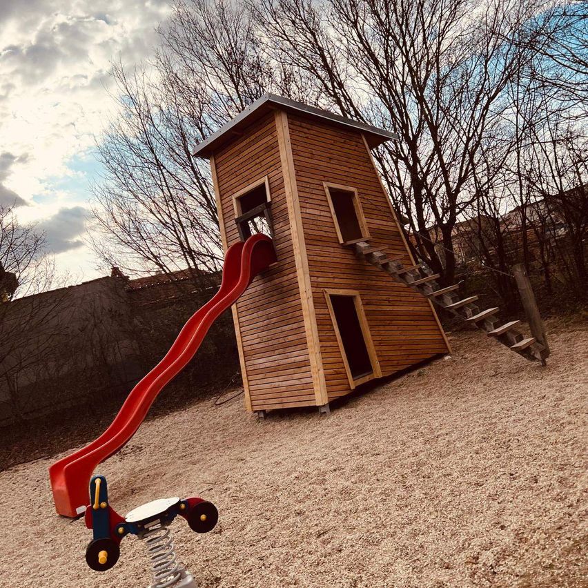 Ein hölzerne Spielhütte mit einer roten Rutsche und einer Spiralfederschaukel befindet sich auf einer sandigen Oberfläche mit Bäumen im Hintergrund.