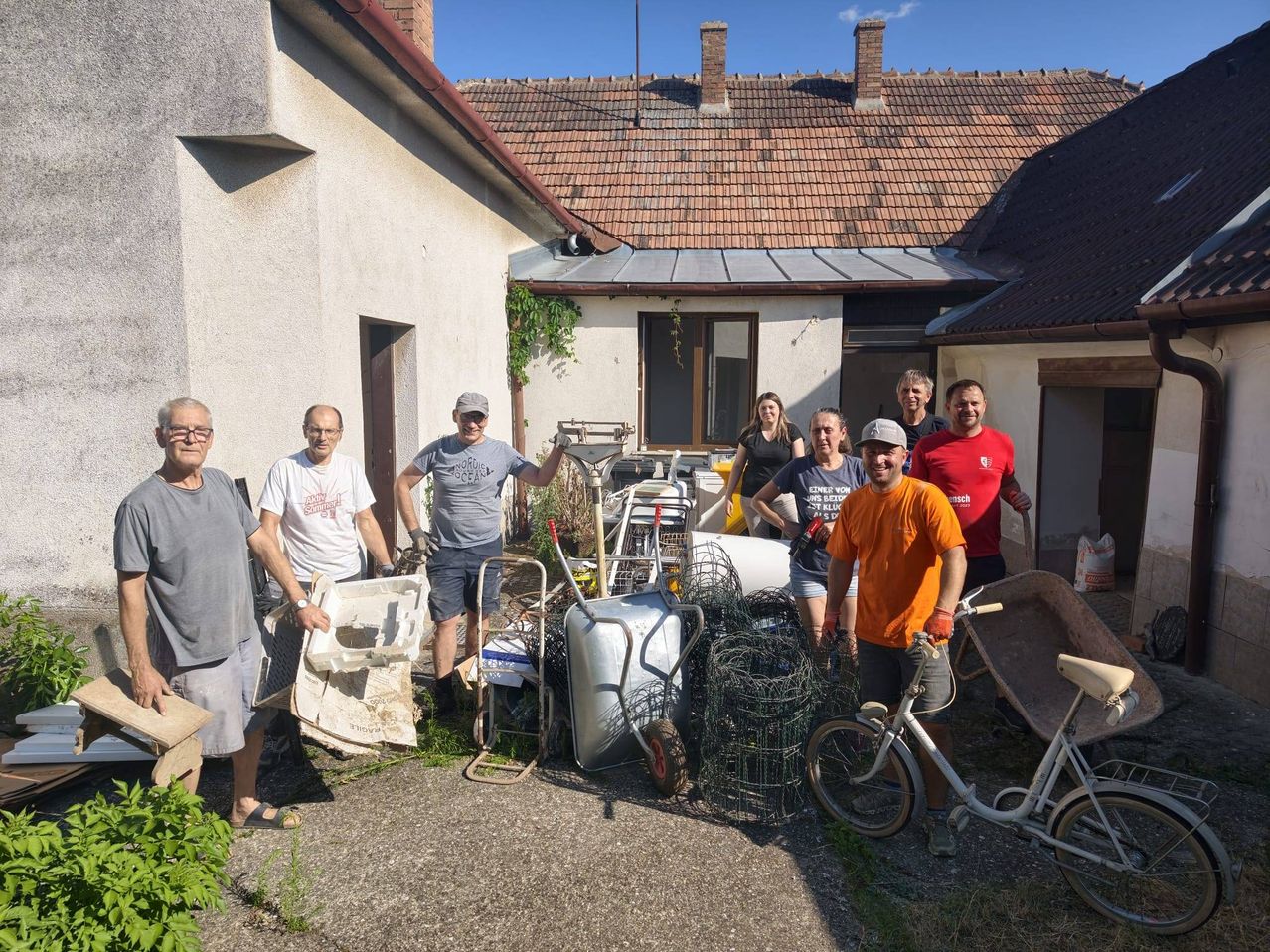 A group of people, including men and women, are gathered in front of a house, posing for a photo. They are holding various items like a wheelbarrow, a white plastic container, and a bicycle. The house has a tiled roof and a chimney.