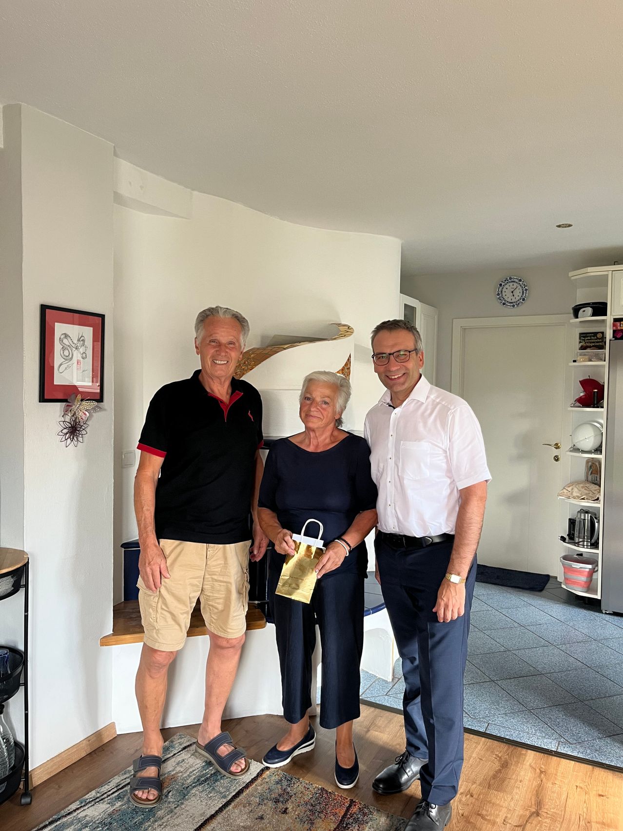 Three people stand together in a room, smiling for a photo. A man in black holds a gift bag, while a woman in blue stands between two men. Behind them is a white wall with a framed picture and a shelf with various items.