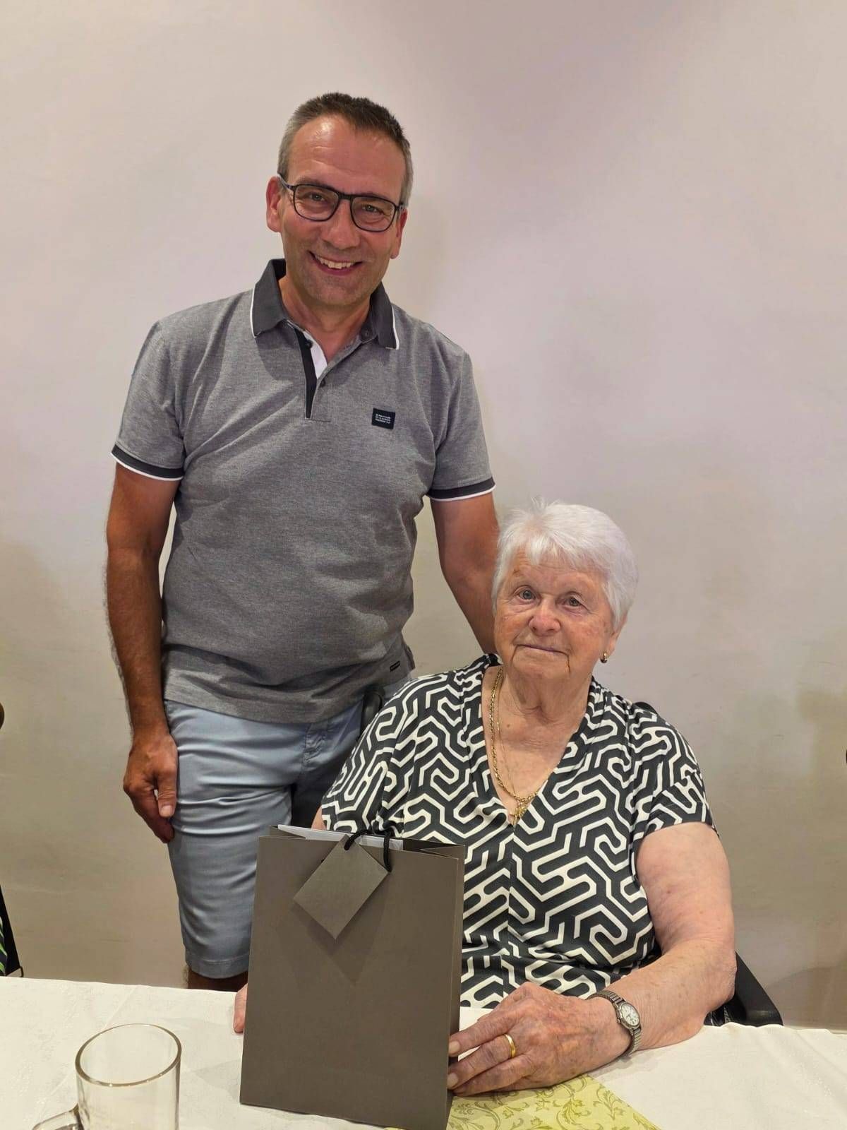 An elderly woman in a patterned blouse sits in a chair, holding a gift bag, while a man in glasses stands behind her, smiling.