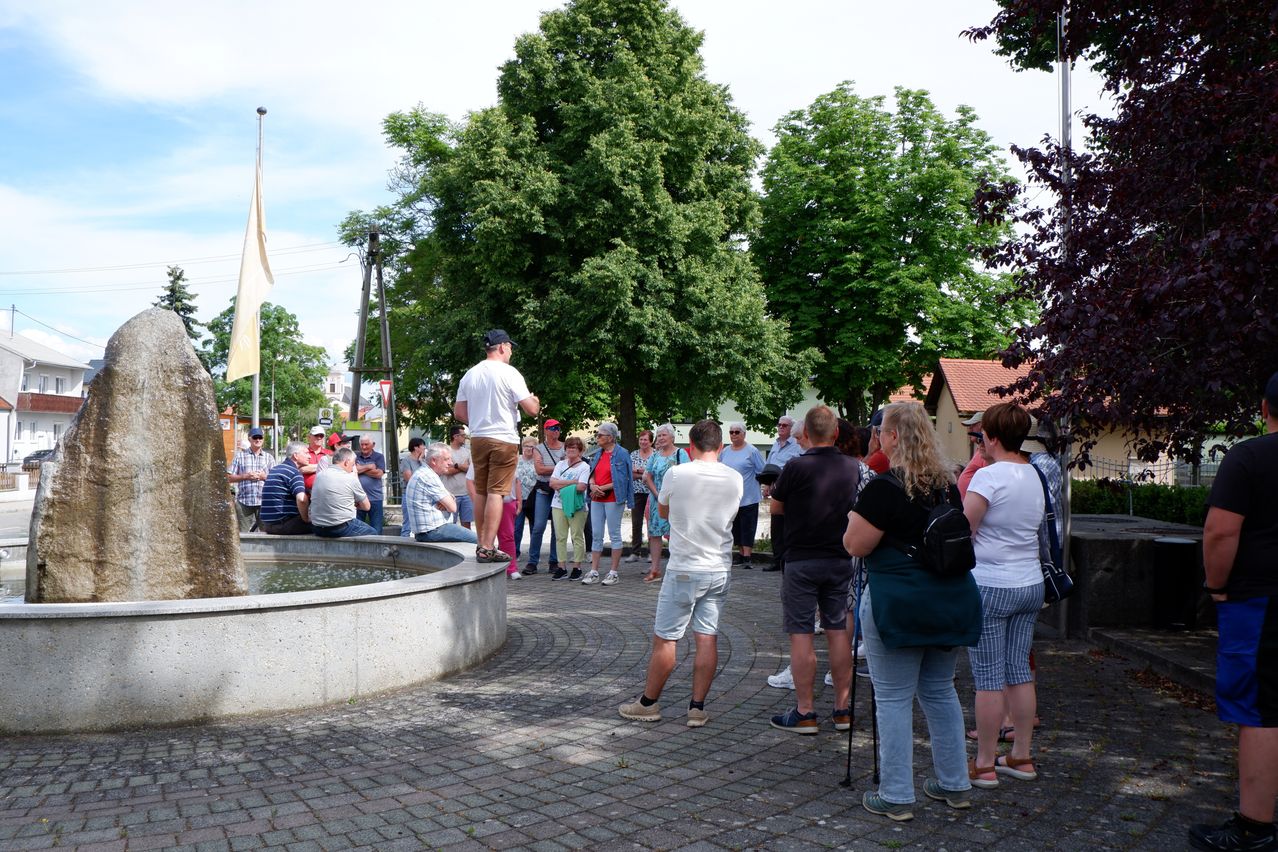 Eine Gruppe von Menschen versammelt sich um einen Brunnen in einem Park. Einige stehen, andere sitzen. Ein Mann steht auf dem Brunnen. Bäume und eine Fahne sind in der Nähe.
