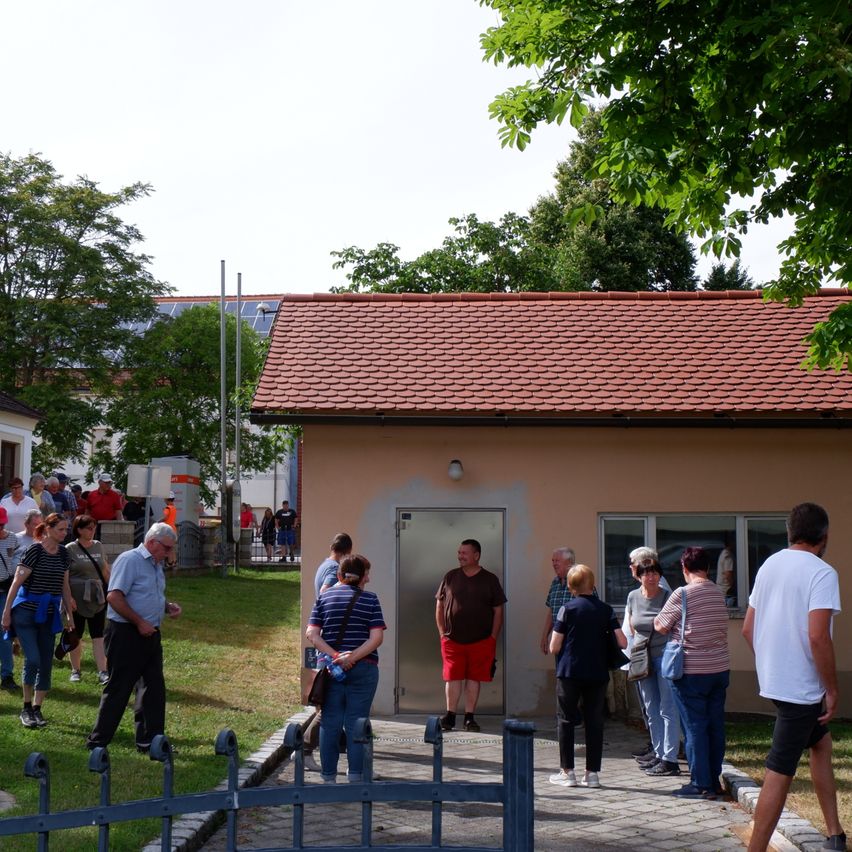 Eine Gruppe von Menschen steht vor einem kleinen Haus mit rotem Dach, umgeben von Grün. Einige unterhalten sich, während andere das Gebäude betrachten. Das Haus hat eine Tür und Fenster.