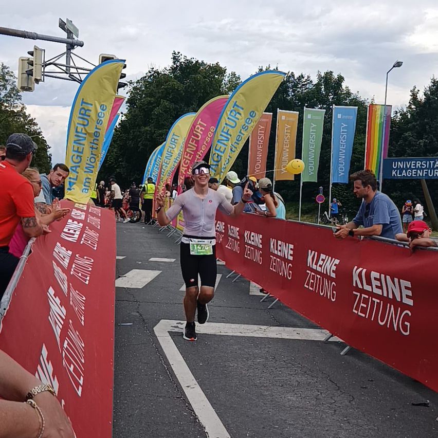 Ein Läufer bei einem Triathlon trägt eine Visierbrille und die Startnummer 1476G. Die Veranstaltung hat bunte Banner und Zuschauer. Die Straße hat eine weiße Linie und eine rote Barriere mit 'Kleine Zeitung' darauf.
