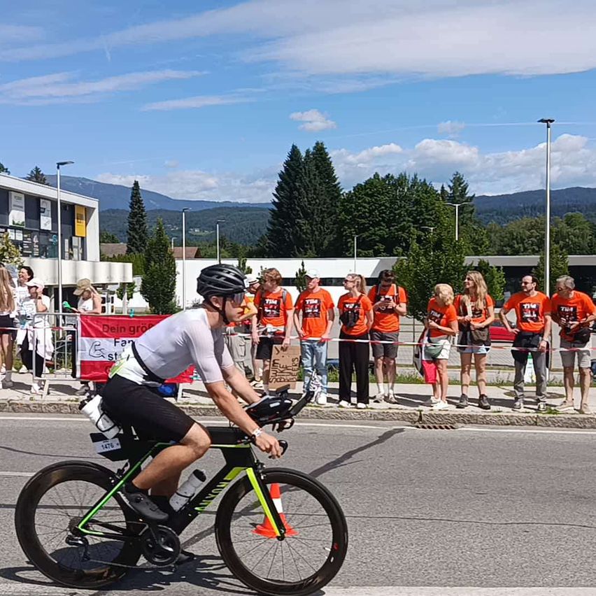 Ein Radfahrer fährt an einer Menschenmenge vorbei an einem sonnigen Tag. Die Menge trägt orangefarbene Shirts und beobachtet den Radfahrer. Der Radfahrer trägt einen Helm und hat Wasserflaschen am Fahrrad befestigt. Bäume und Berge sind im Hintergrund.