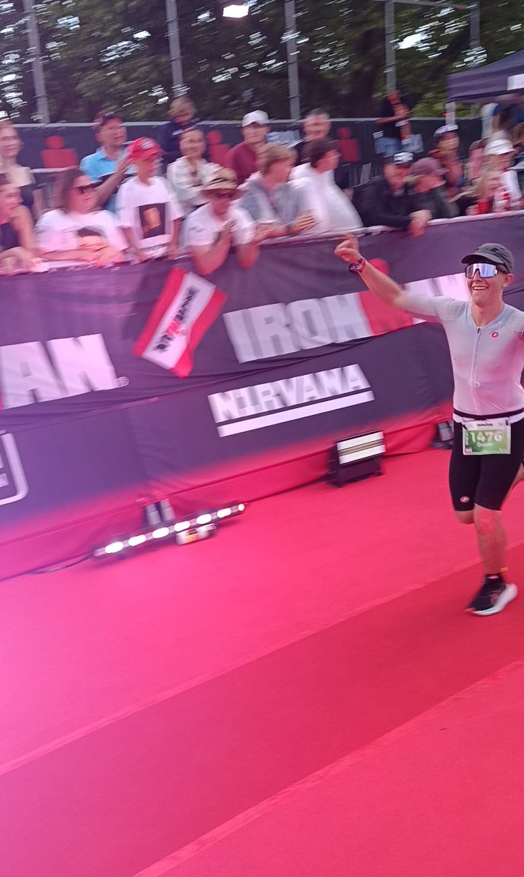 Ein Marathonläufer mit der Startnummer 1476 beendet den Lauf und feiert mit einem erhobenen Arm. Dahinter beobachtet ein Publikum vom Zaun aus.