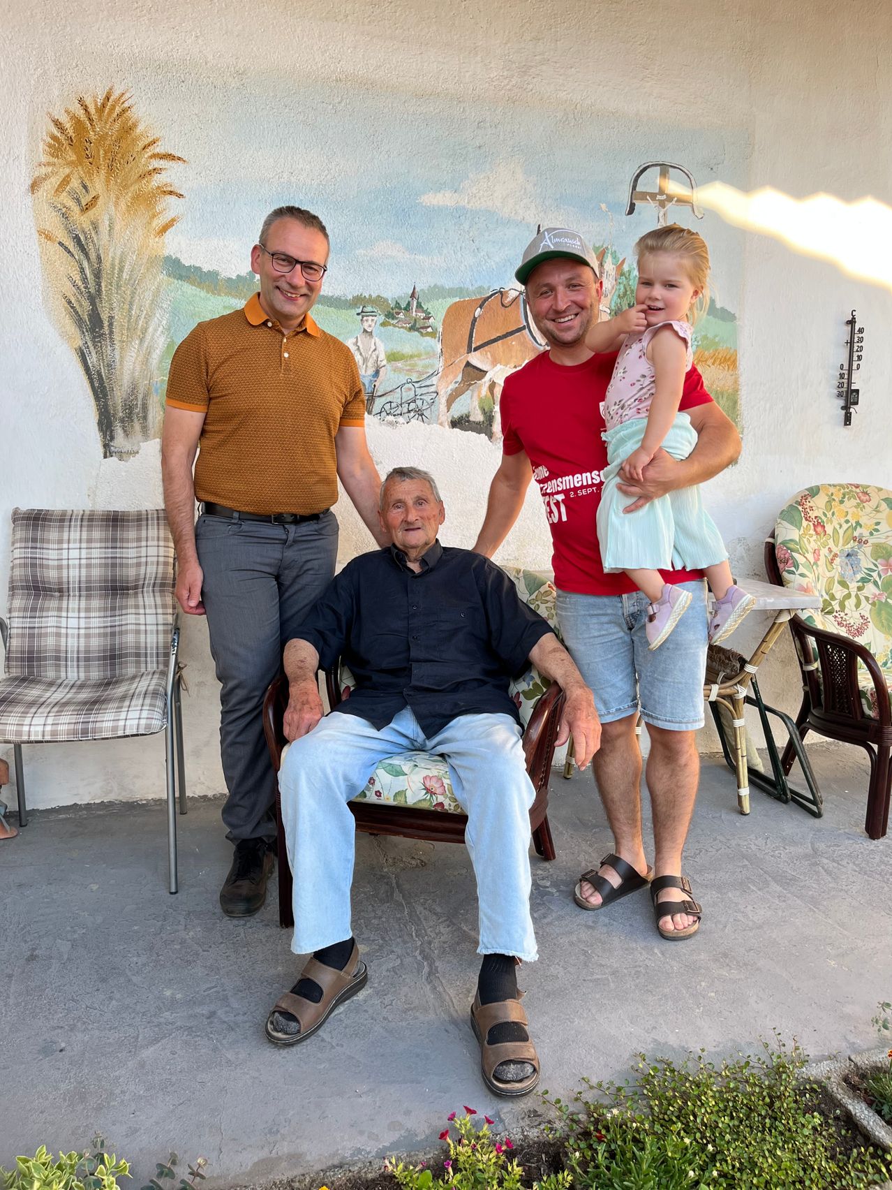 Drei Männer und ein Kind lächeln vor einer Wand mit einem Wandbild und Stühlen. Der ältere Mann sitzt auf einem Stuhl.