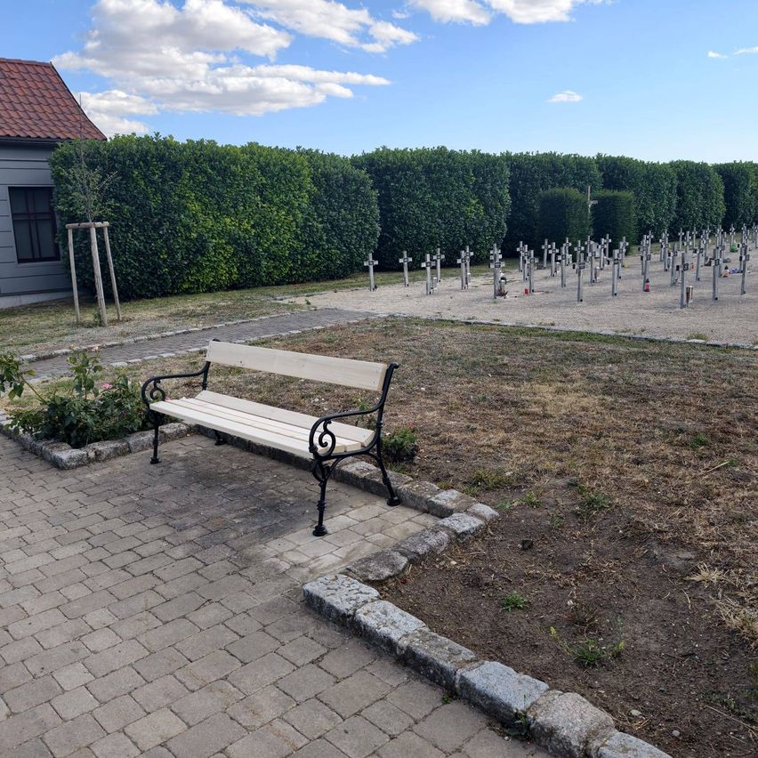 A serene cemetery with a white bench and a row of crosses in the background under a clear blue sky.