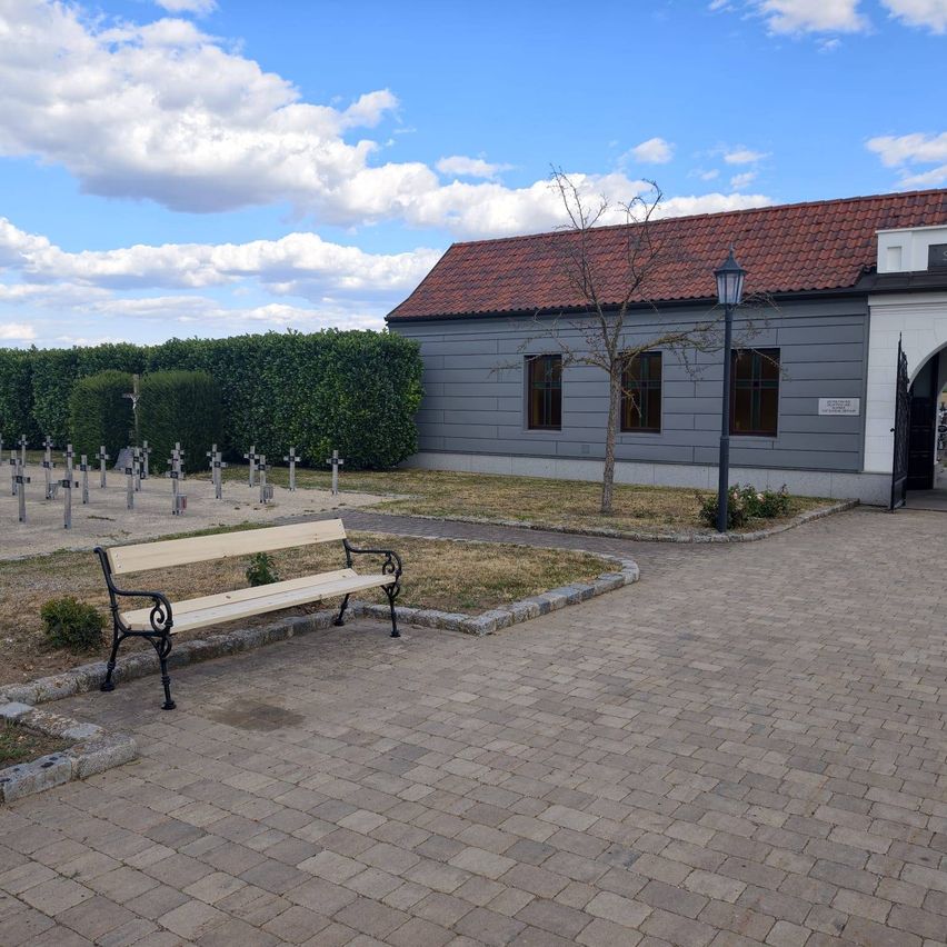 A wooden bench sits in a paved courtyard in front of a building with a red roof. The courtyard is surrounded by a hedge and has several crosses in the background.