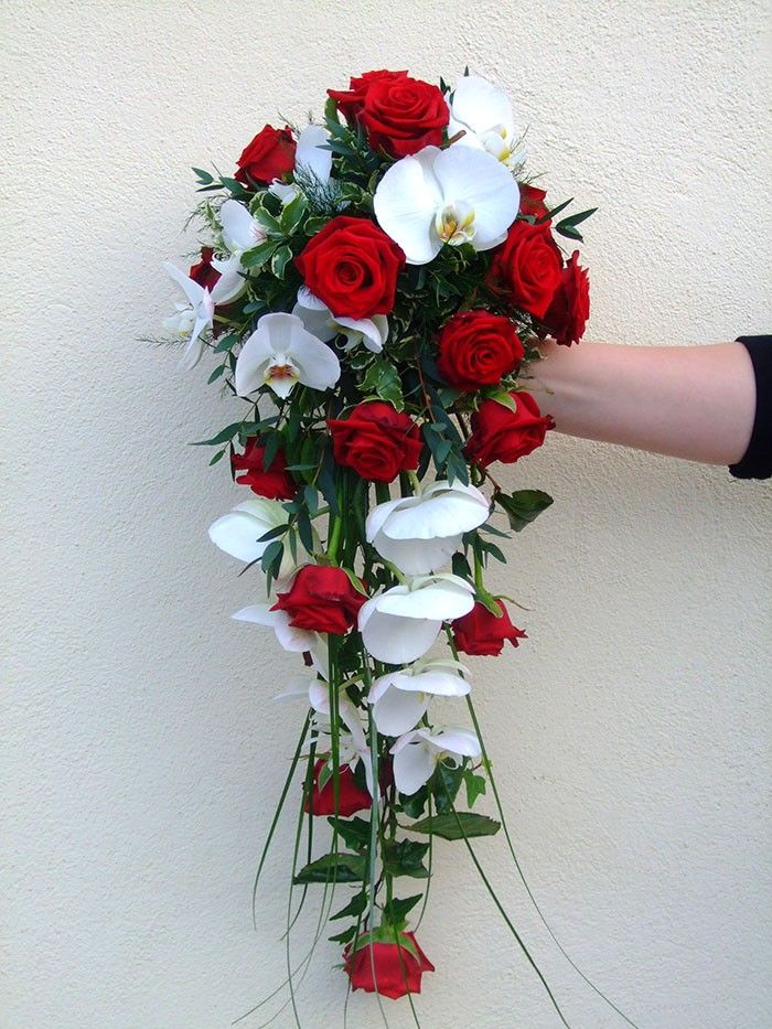 Ein Strauß aus weißen und roten Blumen, einschließlich Orchideen, von einer Hand gegen eine helle Wand gehalten.
