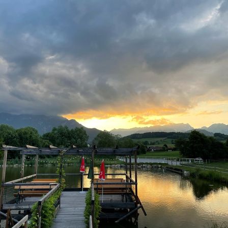 Eine Terrasse am See mit Tischen und roten Sonnenschirmen unter einem bewölkten Himmel mit Bergen im Hintergrund.