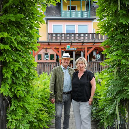 Ein Paar steht lächelnd für die Kamera in einem Garten. Hinter ihnen steht ein farbenfrohes Haus mit Balkon und Schild. Sie sind von viel Grün umgeben.