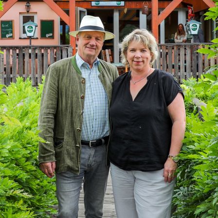 Ein Mann und eine Frau stehen vor einem bunten Haus mit Balkon und Garten. Der Mann trägt einen Hut und eine Jacke, während die Frau ein schwarzes Oberteil und Hosen trägt. Sie lächeln und posieren für ein Foto.