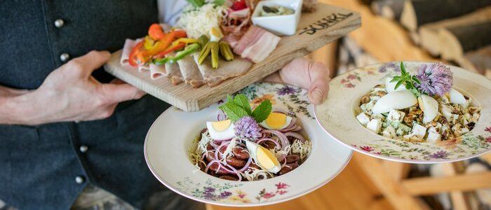 Ein Mann in traditioneller Kleidung hält zwei Teller, einen mit einem Fleischplatten und den anderen mit einem Gericht aus Eiern und Gemüse.